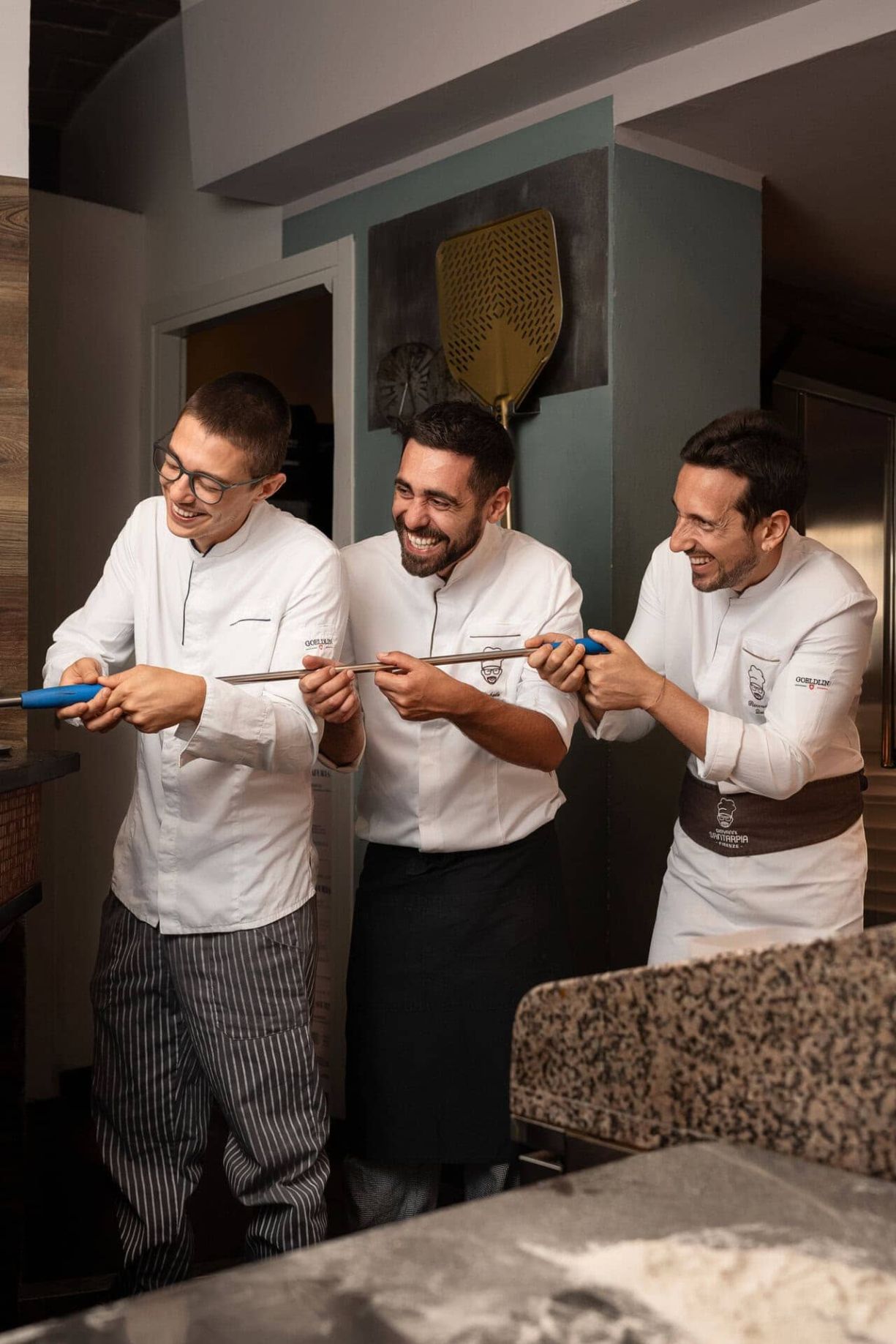 Tre pizzaioli sorridono mentre tengono insieme, in modo scherzoso, un lungo attrezzo da pizza in cucina.